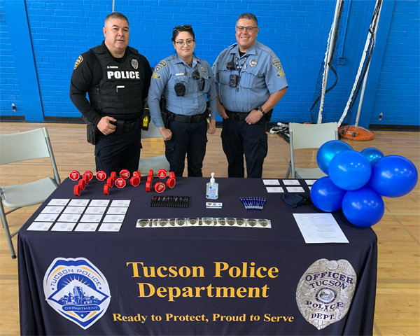 Two CSOs and a police officer, all in uniform, stand behind a recruiting table in a gym.