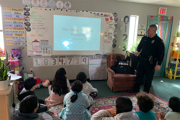 Young children sit on the floor in front of a screen "The Book with No Pictures." A TPD officer in uniform is presenting.