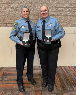 Two smiling female CSOs stand in front of an indoor block wall, displaying their medals.