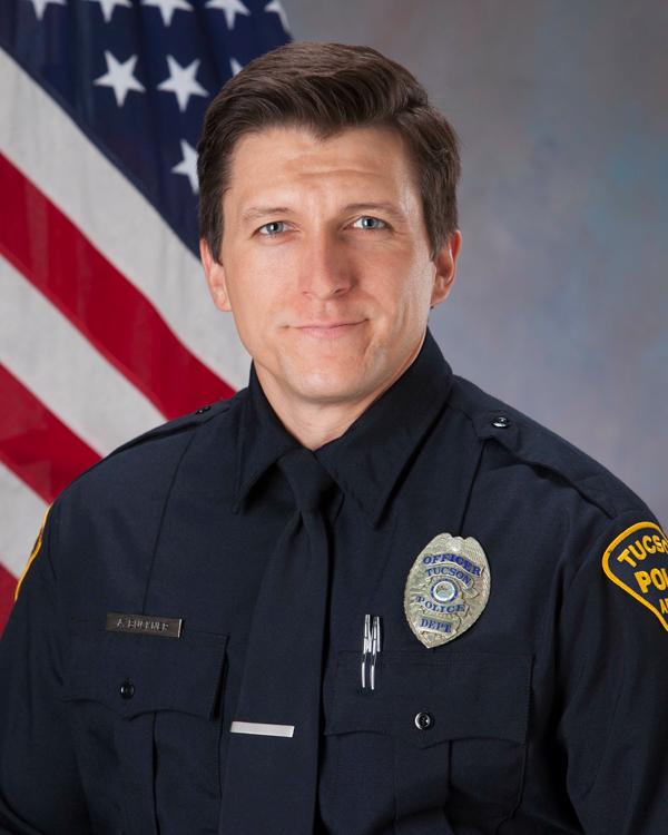 Department photo of a smiling male officer in uniform in front of an American flag.