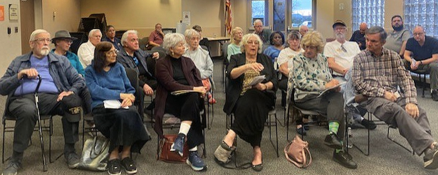 An older audience sits in a public meeting room, an American flag in the background.
