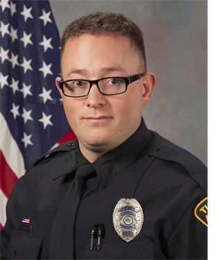 Department photo of male officer in uniform in front of an American flag.