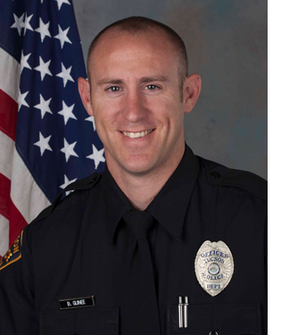 Department photo of smiling male officer in uniform in front of an American flag.