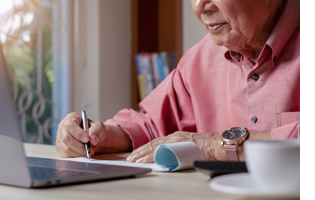 An older man sits at a table at home, writing a check.