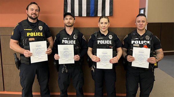 Four officers in uniform stand holding their commendations in front of an American flag with one stripe replaced by a blue line.