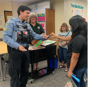 A smiling TPD CSO in uniform hands out school supplies to a student.