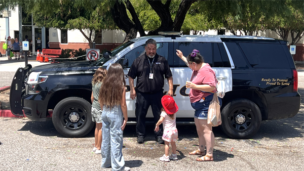 A TPD officer in front of a patrol car interacts with a family with children. A young girl wears a toy firefighter's hat.