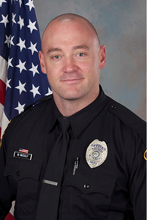 Department photo of officer in uniform in front of an American flag.