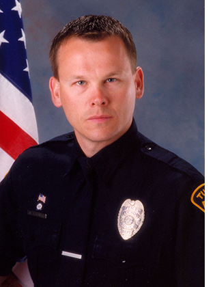 Department photo of TPD officer in uniform in front of an American flag.