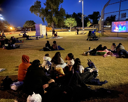 Groups of families sit on blankets on the grass watching a large outdoor movie screen at night.