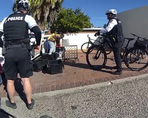 Two officers on bikes contact a male who is bent over by various objects near a vehicle in a residential area.