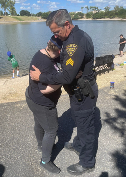 TPD officer in uniform hugs a young person, face away from camera, while other youth are fishing at a small pond in the background.