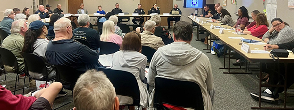 A well-attended meeting with people sitting at long tables against the front and right-hand wall and others sitting in the audience.