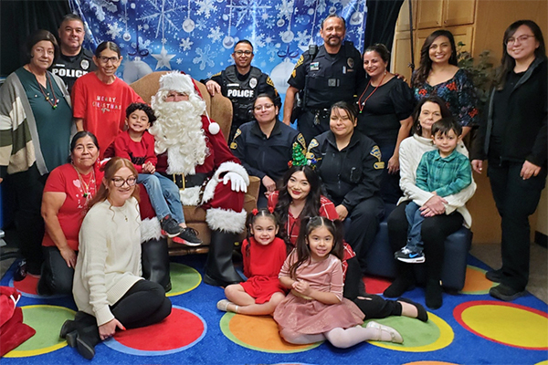 A very tall Santa Claus sits surrounded by children and adults in front of a snowy tableau.