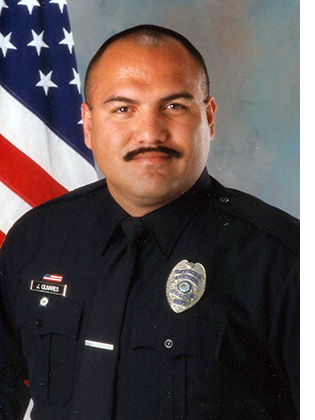 Department photo of male officer in uniform in front of an American flag.