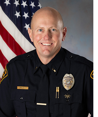 Department photo of male officer in uniform in front of an American flag.