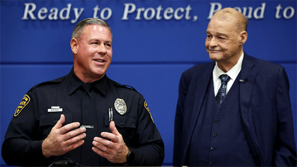 Photo of a male officer in uniform at a podium, an elderly man looks on.