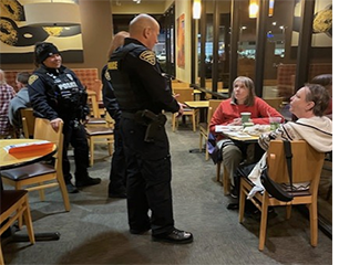 A male officer in uniform stands chatting with customers at a table.