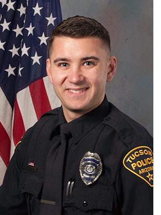 Department photo of a male TPD officer in uniform in front of an American flag.