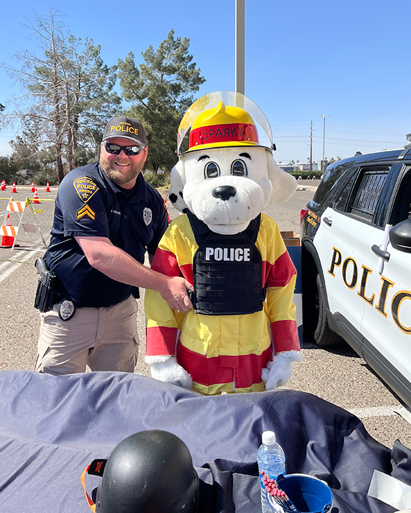 TPD officer gestures toward Tucson Fire mascot "Sparky" (a person in a dog costume), who is wearing a police ballistic vest.