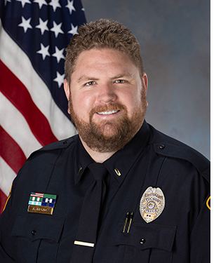 Department photo of a male TPD officer in uniform in front of an American flag.