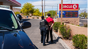 Photo of female TPD officer escorting male suspect in handcuffs. Both are seen from the back. Scene is near a Circle K.
