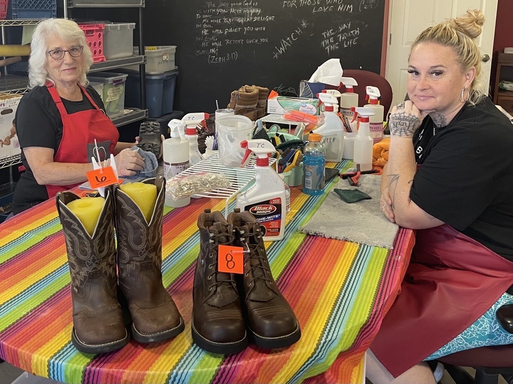 Photo of Staff members Becky and Linette in the cobbler shop with two pairs of freshly cleaned boots.  