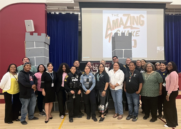 Group photo of TPD members in uniform and school staff in front of the stage in a school auditorium.