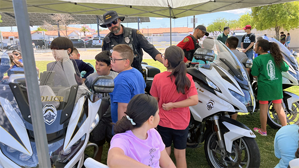 Children under a pop-up tent looking at police motorcycles with police officers in SWAT and academy uniforms.
