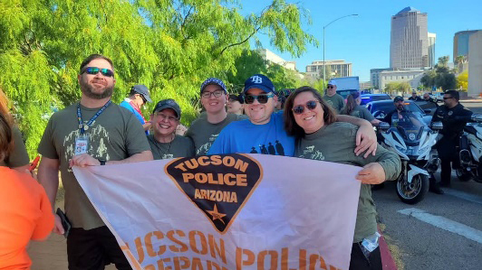 TPD members wearing Torch Run T-shirts hold a TPD banner. TPD motorcycles are in the background. 