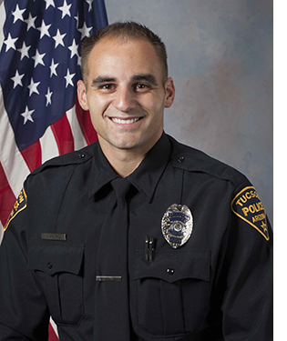Department photo of male officer in uniform in front of an American flag.