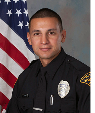 Department photo of male officer in uniform in front of an American flag.