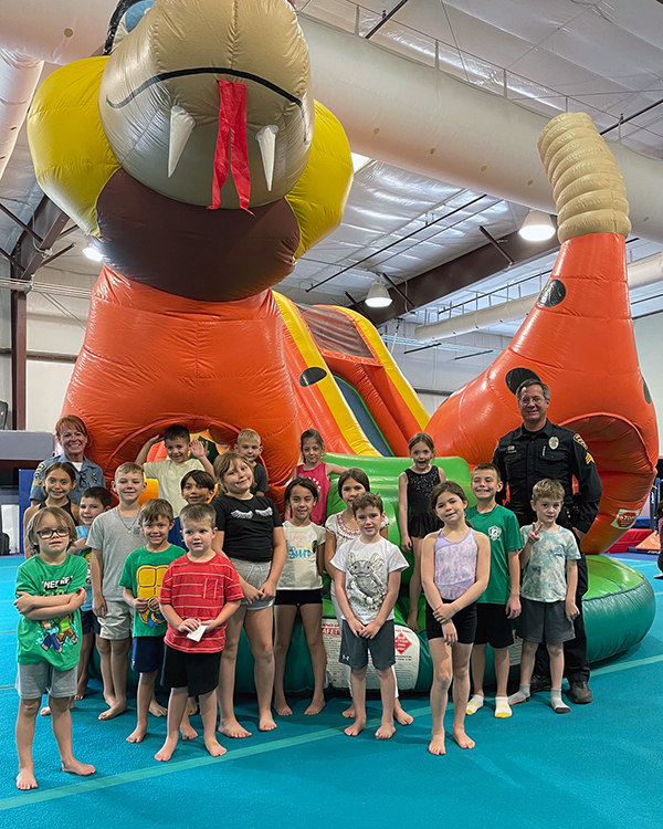 Barefoot children and a uniformed officer stand in a large gym with a huge inflatable snake, like a bouncy castle, in the background.