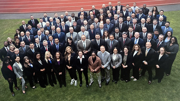 Group photo taken from a drone of a large group of people in suits standing on grass in front of an outdoor track, looking up at the drone.