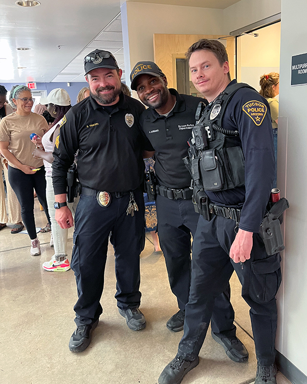Three smiling officers in uniform stand in a doorway. Community members are seen in the background.