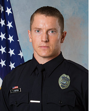 Department photo of male officer in uniform in front of an American flag.