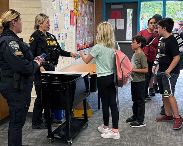 Two female officers in uniform hand out backpacks in a school hallway.