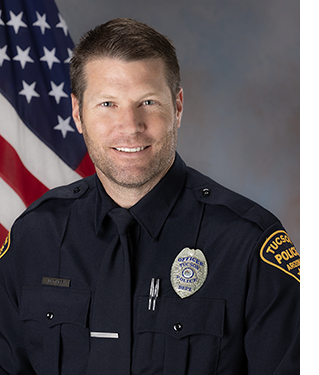Department photo of a smiling male TPD officer in uniform in front of an American flag.