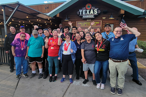 A large group of smiling people, some in uniform, some waving, stand in front of Texas Roadhouse.
