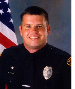 Department photo of smiling male officer in uniform in front of an American flag.