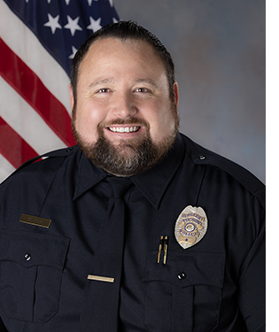 Department portrait of a smiling male officer in uniform in front of an American flag.