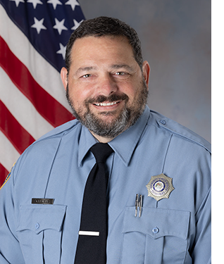 Department portrait of smiling male CSO in uniform in front of an American flag.