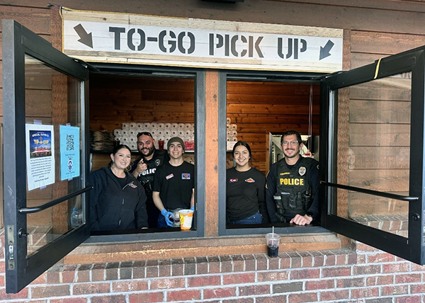 Group photo of uniformed officers at an open service window looking outside. Above the window reads, "To-Go Pick Up."