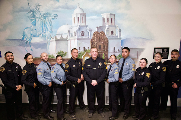 Group photo of officers and CSOs in front of a mural depicting Mission San Xavier del Bac and a ghostly Father Kino on horseback.