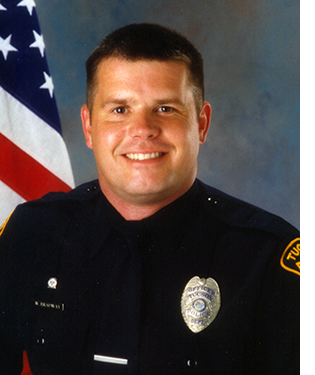 Department photo of smiling male officer in front of American flag.
