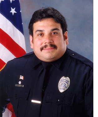 Department photo of male officer in uniform in front of an American flag.