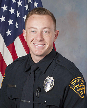 Department photo of smiling male officer in front of an American flag.