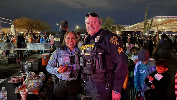 A TPD officer and a CSO in uniform smile for the camera at a nighttime Halloween event. Many kids and families are seen queuing up for candy..