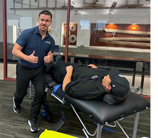 A uniformed male TPD member, fully clothed, lies on a massage table, facing up. Another male Stretch Zone staff gives a thumbs up to the camera.