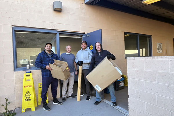Four guys in work clothes stand at a building entrance with large empty cardboard boxes.